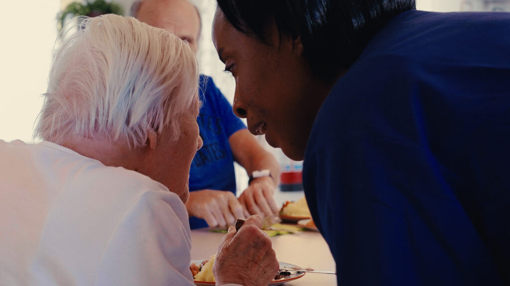 en äldre man och kvinna sitter vid ett matbord. En yngre kvinna i vårdkläder samtalar med dem.