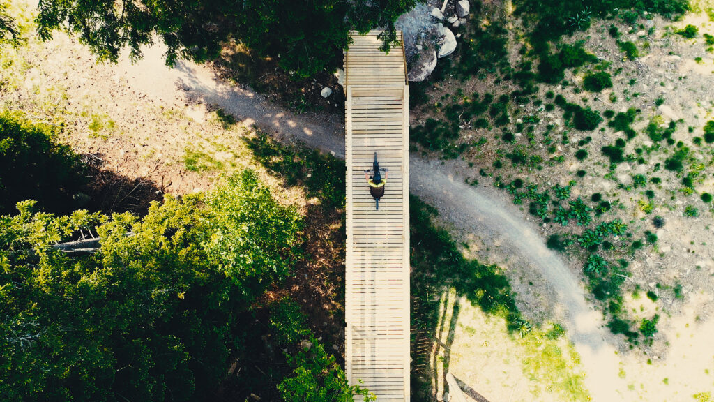 Drönarbild från ovan på en cyklist på en träbro över en grusstig med omkringliggande skog.