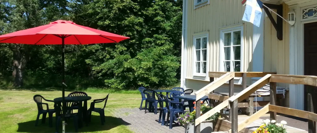 Trädgårdsmöbler och ett parasoll på en gräsmatte med lummig bakgrund. Trappen till ett hus med servering finns till höger.