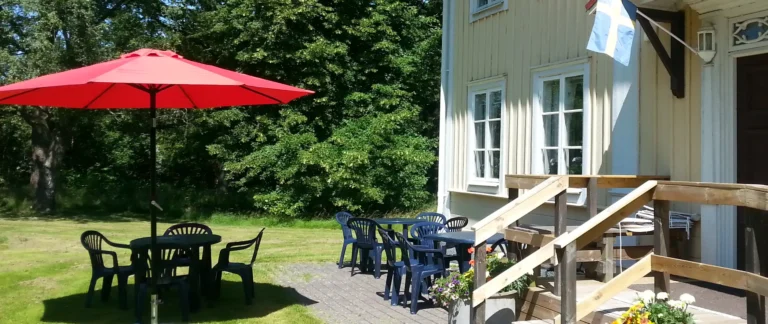 Trädgårdsmöbler och ett parasoll på en gräsmatte med lummig bakgrund. Trappen till ett hus med servering finns till höger.