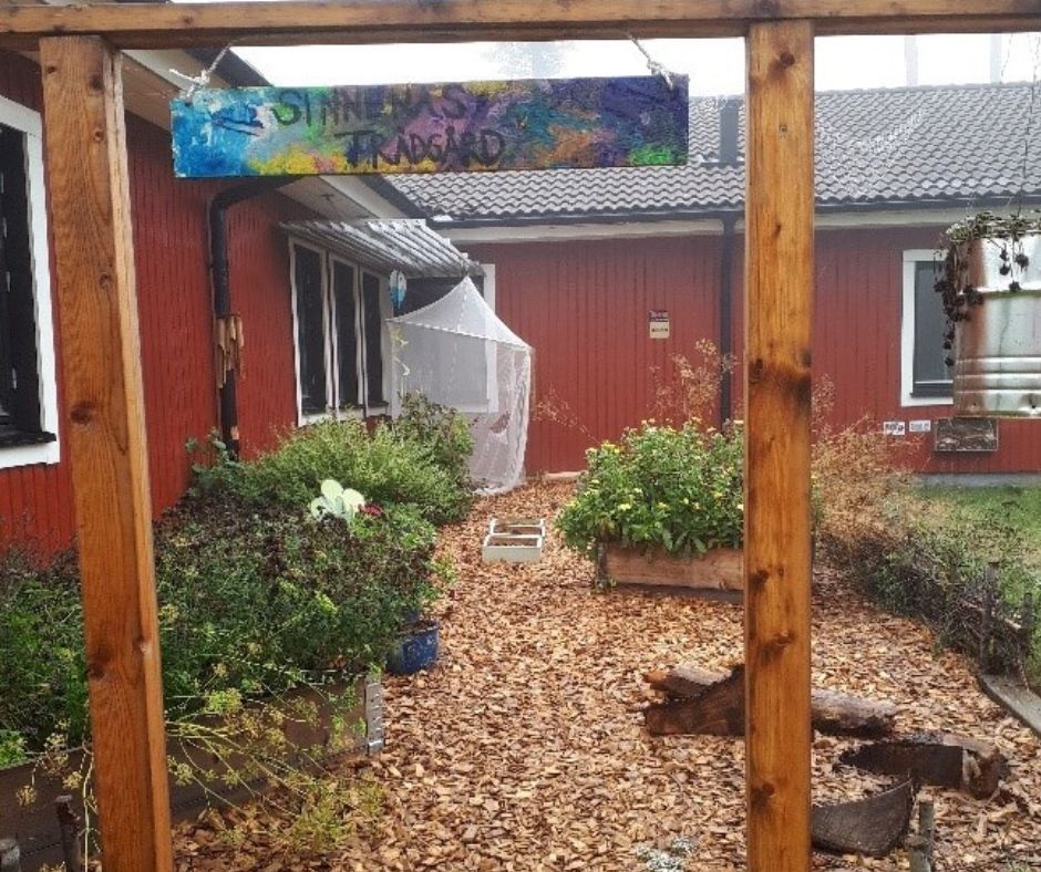 Gång i gårdsyta med träflis och odlingar. Intill ett rött trähus. Portal med färgglad skylt som säger "sinnernas trädgård"