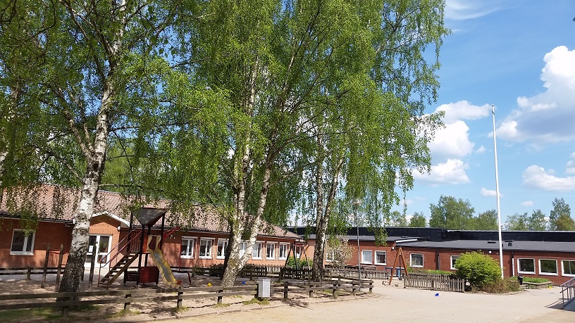 Skolbyggnader i tegel. Ytor omgärdade av staket med grönskande träd och lekredskap.