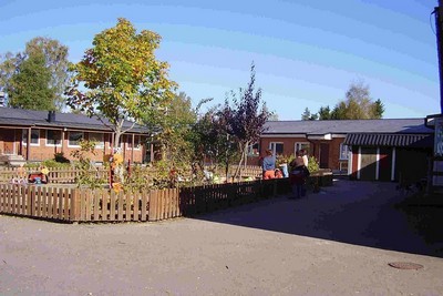 Förskolebyggnad i orange tegel med en lummig innergård. Staket och lekredskap.