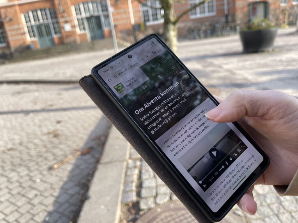 En hand som håller i en mobiltelefon. På skärmen visas en sida med titeln Om Alvesta kommun