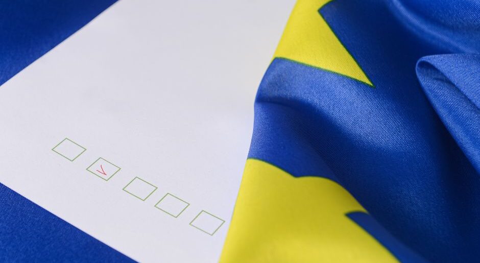 Ett röstkort med en bock i en ruta ligger på ett blått underlag. EU:s blåa flagga med gula stjärnor ligger vid ena kanten.