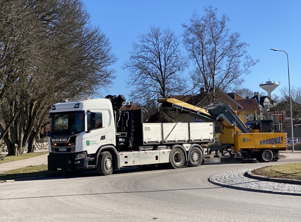 Maskin sopar en rondell från grus och tömmer samtidigt i en lastbil som kör framför.