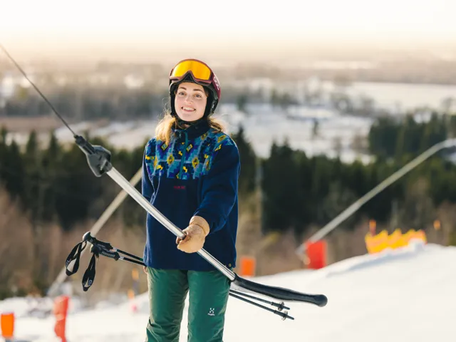 Närbild på kvinna med skidglasögon upphissade på  hjälmen. Åker lift upp för backe. 