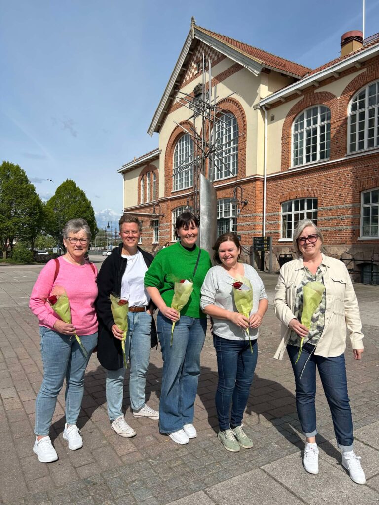 5 kvinnor står utanför ett stationshus. Alla ha varsin blombukett i handen,