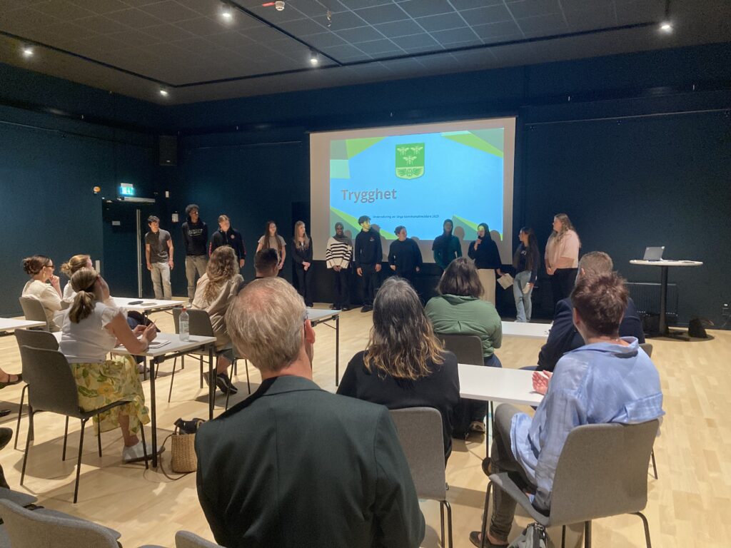 En grupp unga står framför en PPT-presentation på en stor skärm. Åhörare sitter framför.