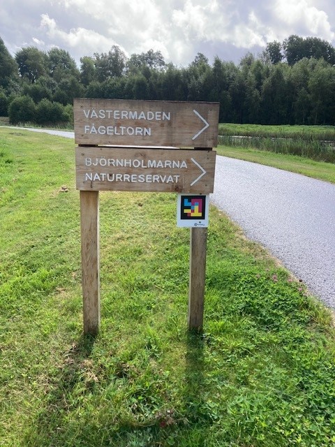 En vägvisare med pilar mot Västermaden fågeltorn och Björnholmarna naturreservat. Under skylten sitter en färgstark skylt med en navilens-kod.