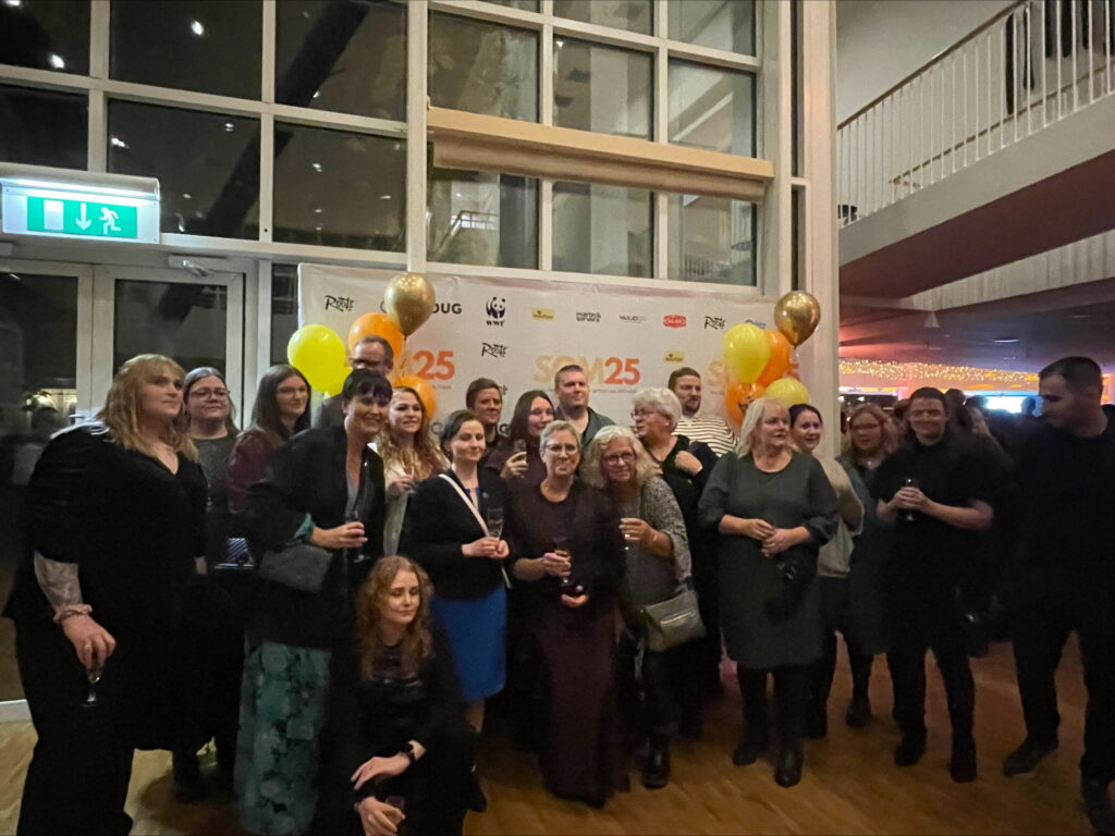 Flera personer står uppställda för ett gruppfoto framför en ballongprydd monter där det står SOM2025 på.