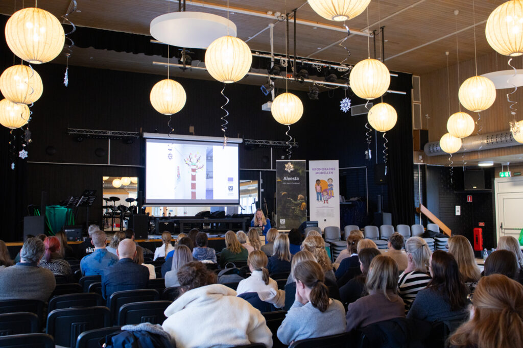 en stor grupp människor sitter i ett mörk rum och tittar på en föreläsning.