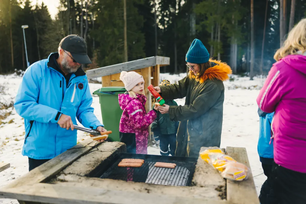Två vuxna, en man och en kvinna, samt ett barn står vid grillplats. mannen lägger en korv i ett bröd. Kvinnan syns hälla ketchup på en korv. Det är vinter.