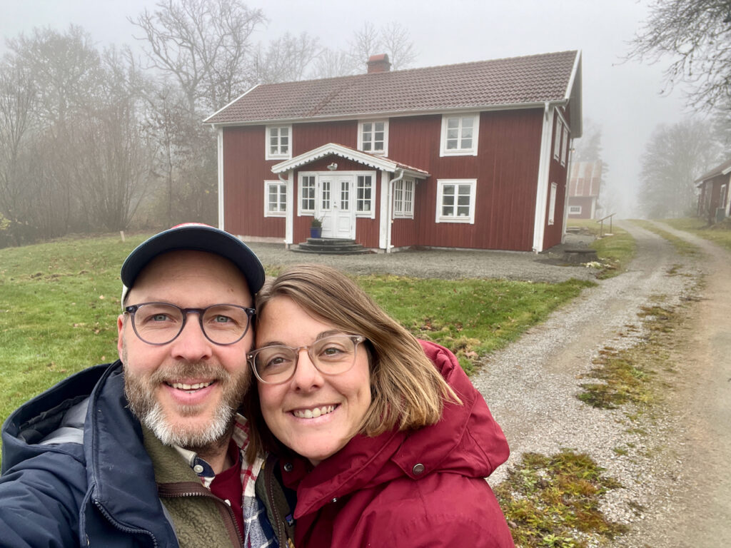 En man och kvinna håller om varandra framför sitt röda hus på landsbygden.
