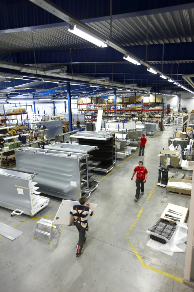 Stor industrilokal. På sidorna finns höga lagerhyllor och i mitten syns tre personer på håll.