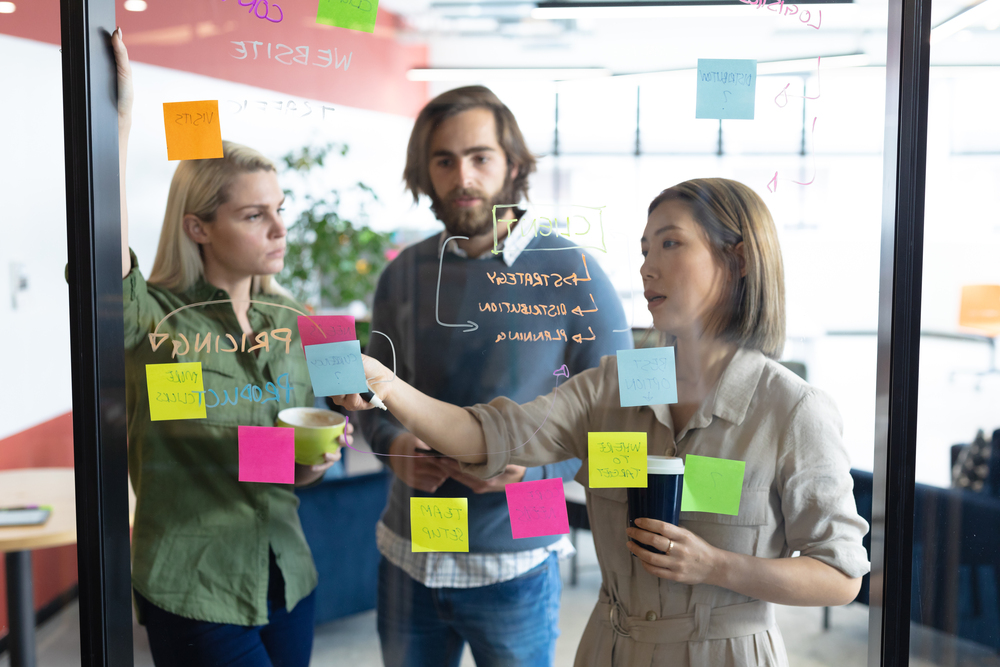 Tre personer står framför en genomskinlig tavla, med text och post-its.