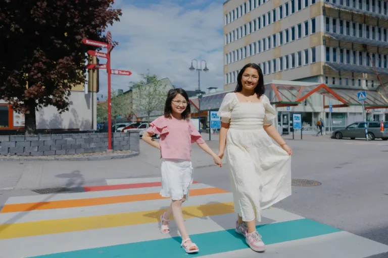 Två flickor som går över ett regnbågsfärgat övergångsställe med Storgatan och kommunhuset i Alvesta i bakgrunden.